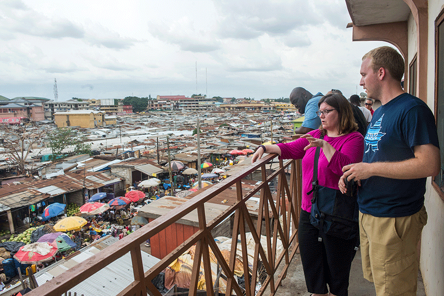 Adult male in navy Indiana State University t-shirt stands next to shorter adult woman in pink top on balcony overlooking a city in another country.