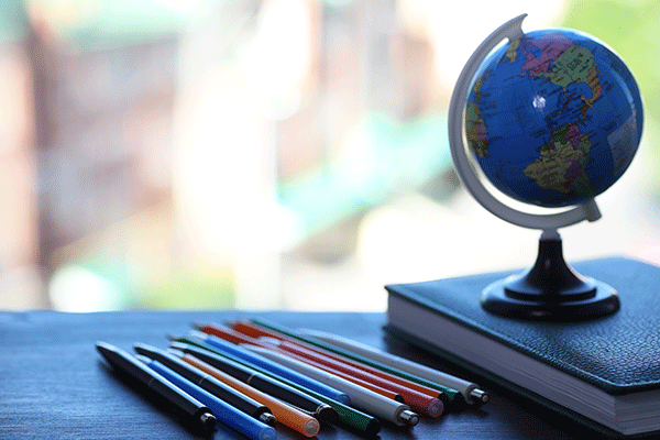 A globe sits on top of a book on a desktop with colored pencils beside the book